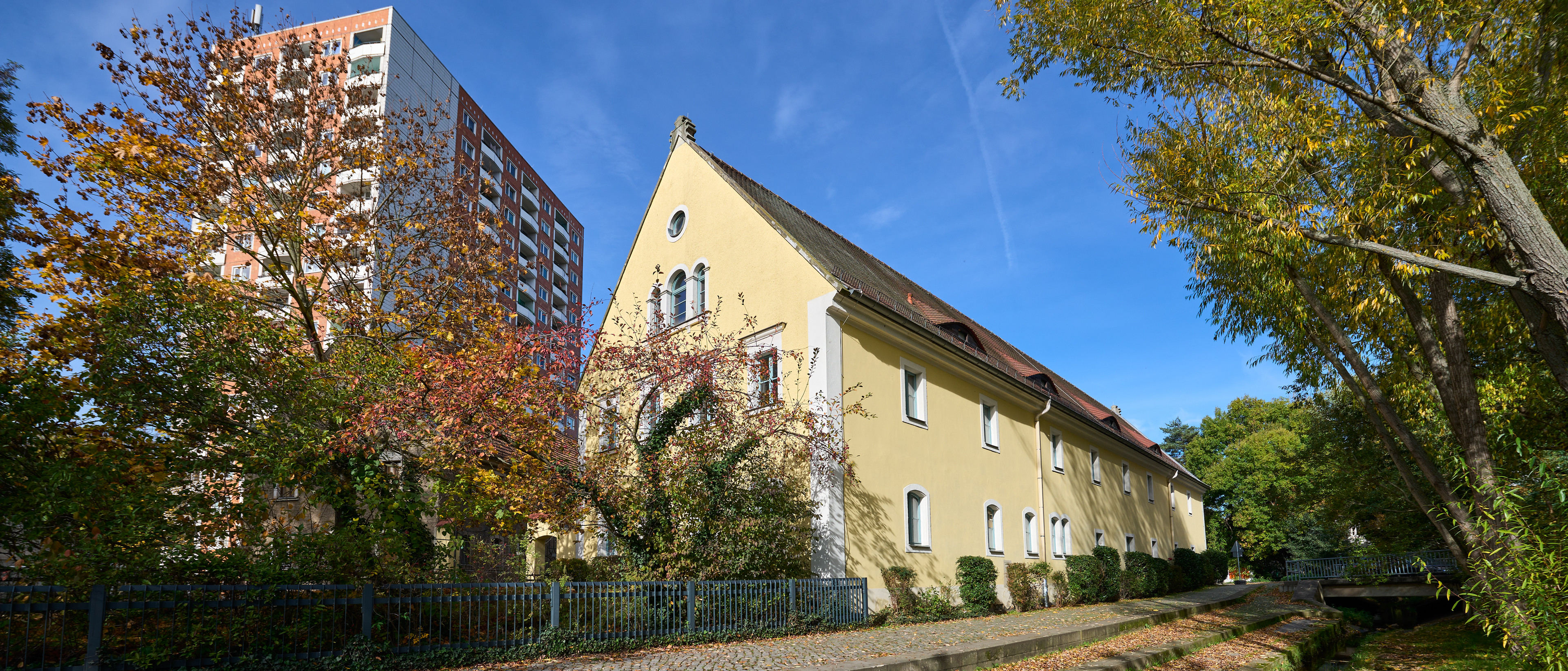 KI generiert: Das Bild zeigt ein gelbes, historisches Gebäude neben einem modernen Hochhaus unter einem klaren blauen Himmel. Im Vordergrund sind herbstliche Bäume und ein schmaler Weg zu sehen.