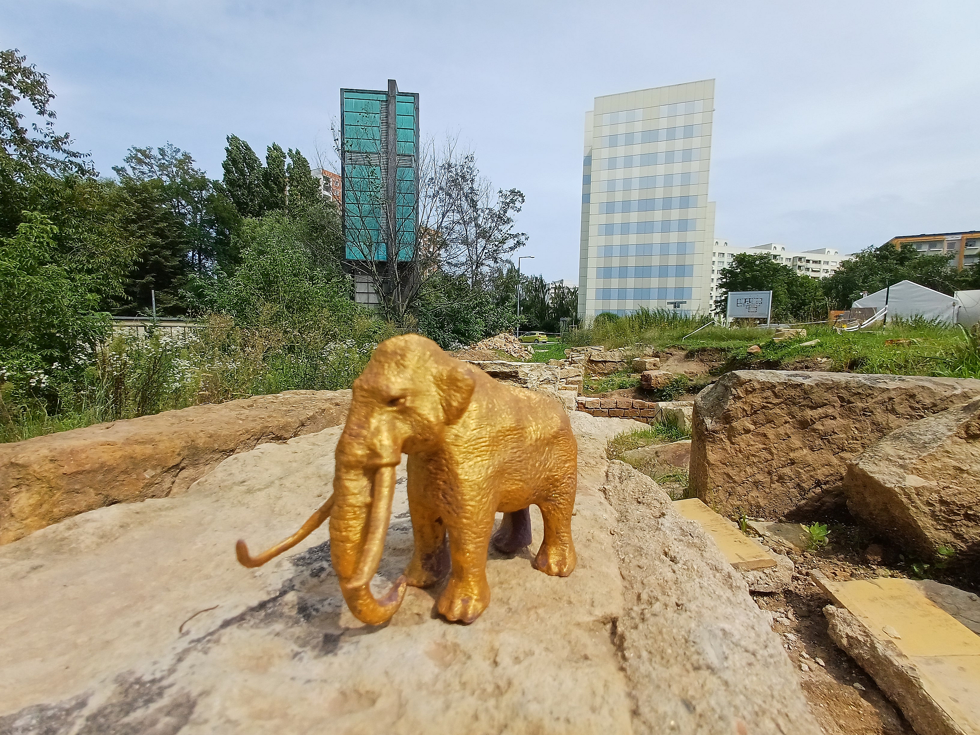 KI generiert: Im Vordergrund des Bildes steht eine goldene Elefantenfigur auf einem steinigen Untergrund, während im Hintergrund moderne Bürogebäude und grüne Vegetation sichtbar sind.