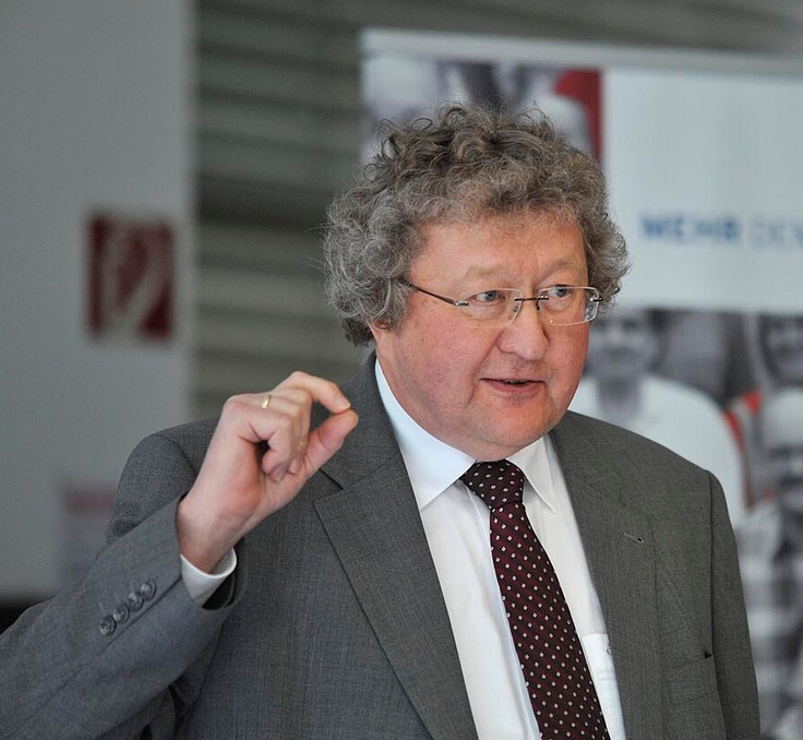 KI generiert: Ein Mann in einem Anzug hält einen Vortrag und gestikuliert leicht mit seiner Hand. Im Hintergrund sind verschwommene Bildausschnitte von Menschen und Text erkennbar.