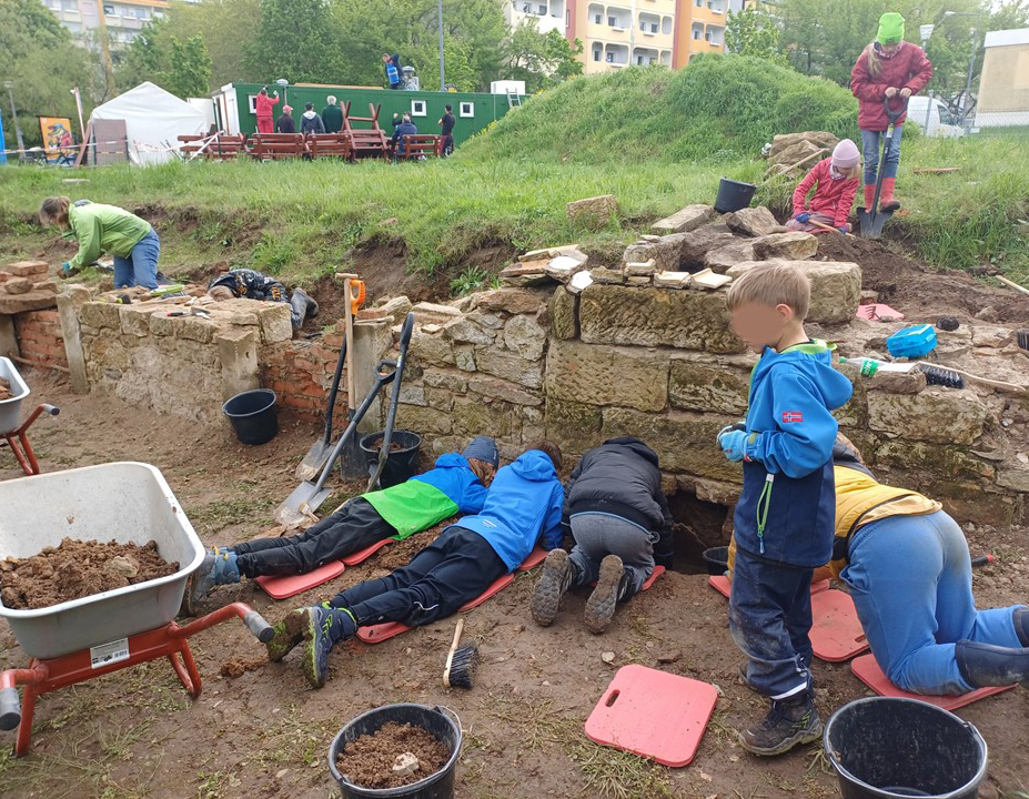 KI generiert: Das Bild zeigt mehrere Kinder und Erwachsene, die an einer archäologischen Ausgrabungsstelle arbeiten, umgeben von Schaufeln, Schubkarren und Erde. Die Szene spielt sich in einem mit Gras bewachsenen Bereich ab, wo die Menschen in einer Grube nach Artefakten suchen.