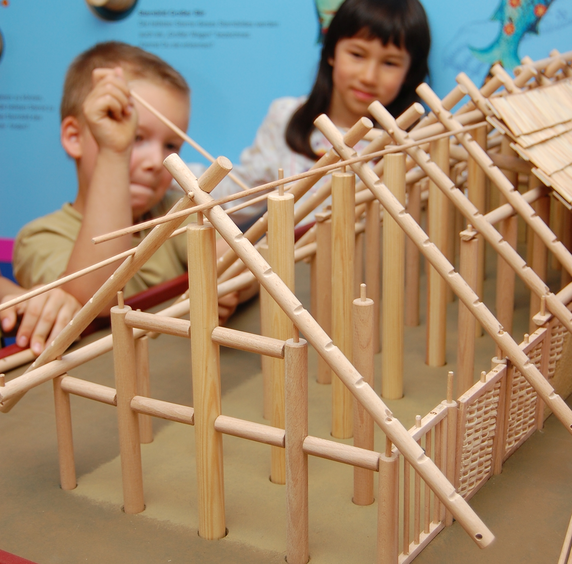 KI generiert: Das Bild zeigt zwei Kinder, die mit einer Holzbaukonstruktion spielen und diese zusammenbauen. Der Fokus liegt auf der teilweise fertig gestellten Struktur und den Kindern im Hintergrund.