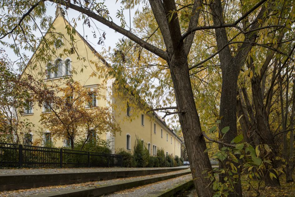 KI generiert: Das Bild zeigt ein langes, gelbes Gebäude mit Giebel und mehreren Fenstern, das von herbstlich gefärbten Bäumen umgeben ist. Im Vordergrund sind ein Weg und ein Wasserlauf zu sehen.