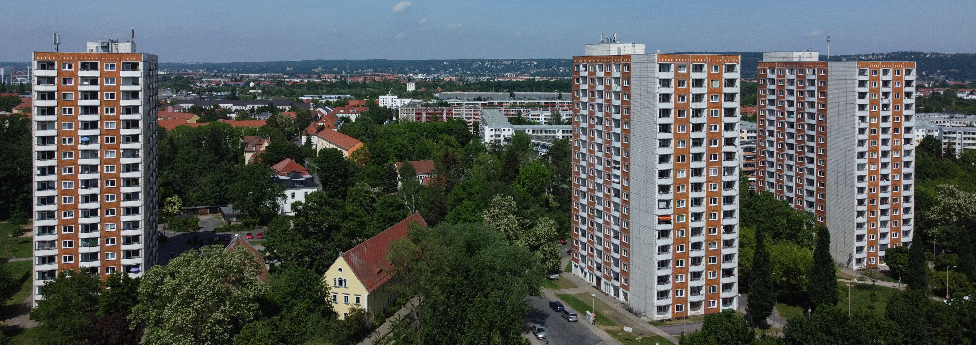 KI generiert: Das Bild zeigt eine städtische Landschaft mit drei hohen Wohngebäuden, die von Bäumen und kleineren Gebäuden umgeben sind. Im Hintergrund sind weitere Stadtteile und Hügel zu erkennen.