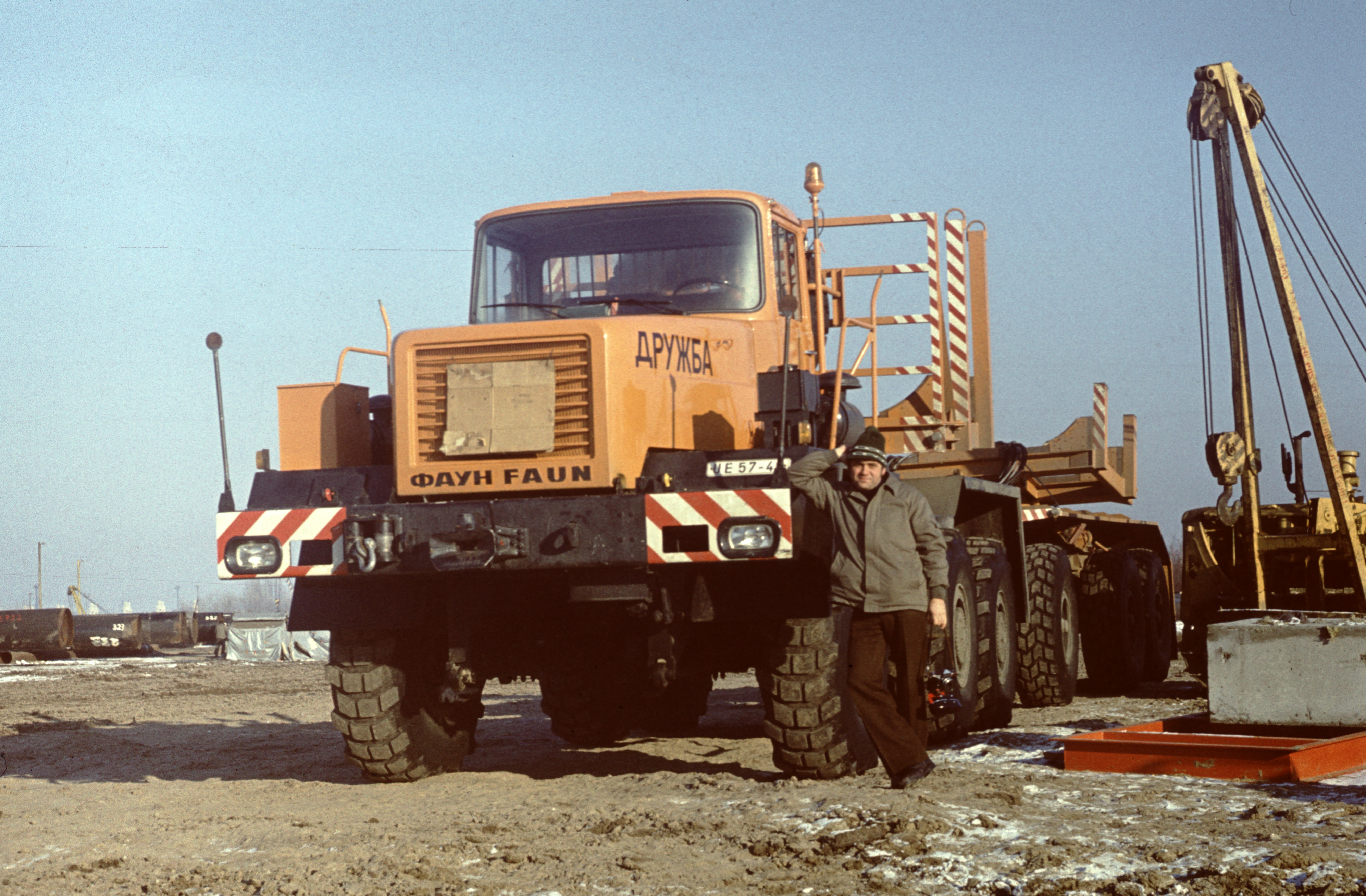 KI generiert: Das Bild zeigt einen großen, orangefarbenen Schwerlast-Lkw von FAUN auf einer Baustelle. Eine Person steht neben dem Fahrzeug und lehnt sich an die Vorderseite.