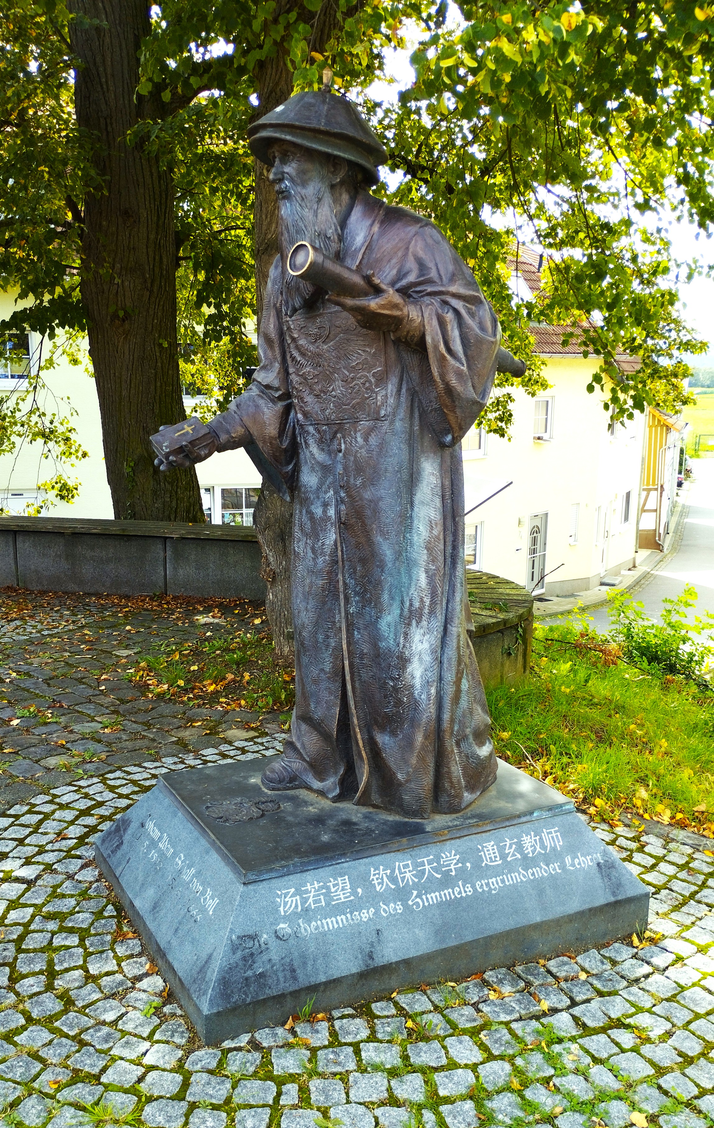 KI generiert: Das Bild zeigt eine Bronzestatue eines Mannes in traditioneller Kleidung, der ein Teleskop hält. Die Statue steht auf einem steinernen Sockel mit Gravuren und befindet sich in einem parkähnlichen Umfeld.