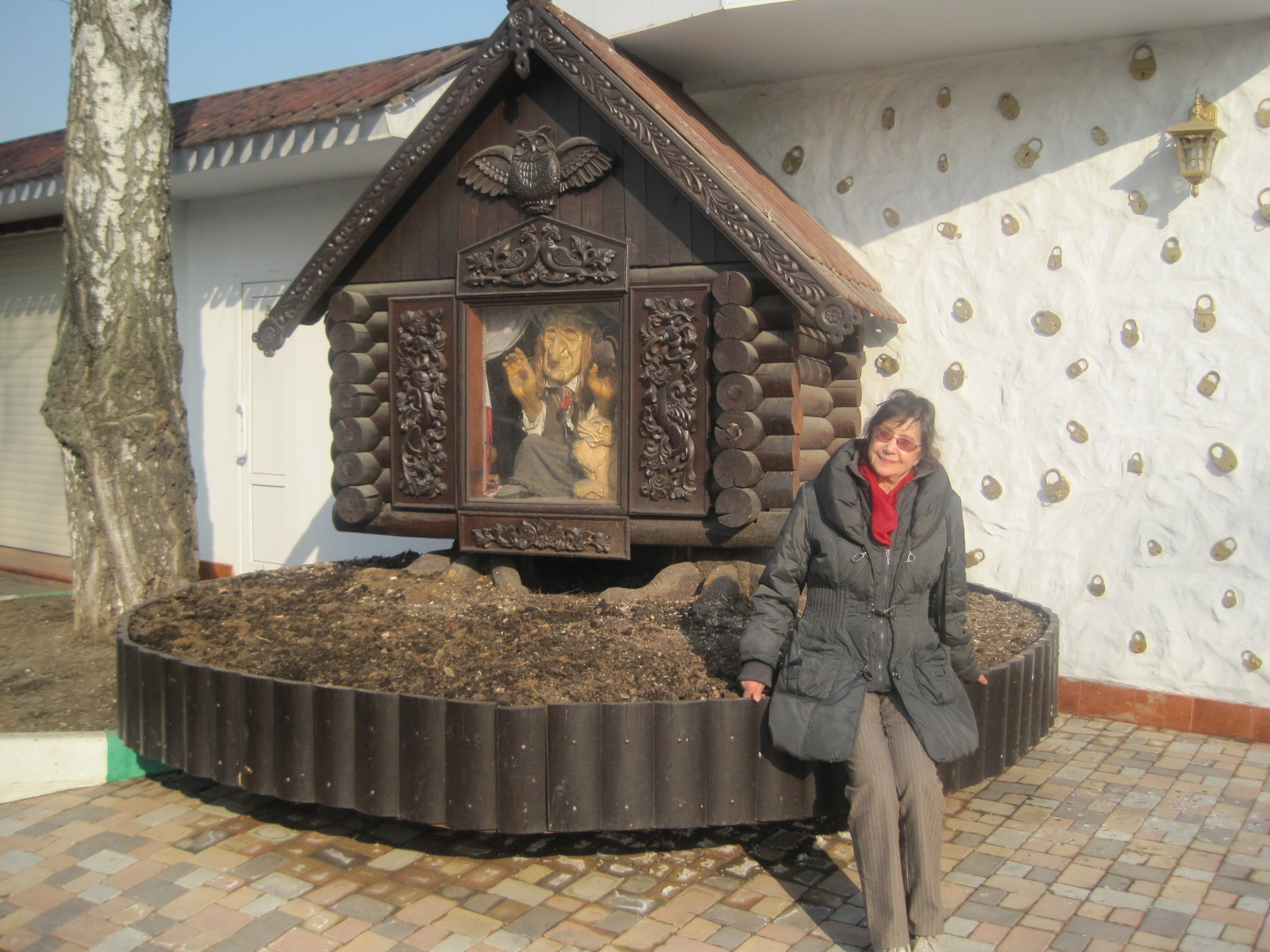 AI generated: A person stands next to a small, decorative wooden house, which is skilfully carved and rests on a raised base. Figures or dolls can be recognised inside the house.