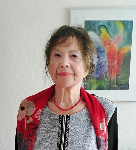 KI generiert: Das Bild zeigt eine ältere Frau, die vor einem farbenfrohen, abstrakten Gemälde an der Wand steht. Sie trägt ein graues Kleid mit einem roten Schal und lächelt leicht.