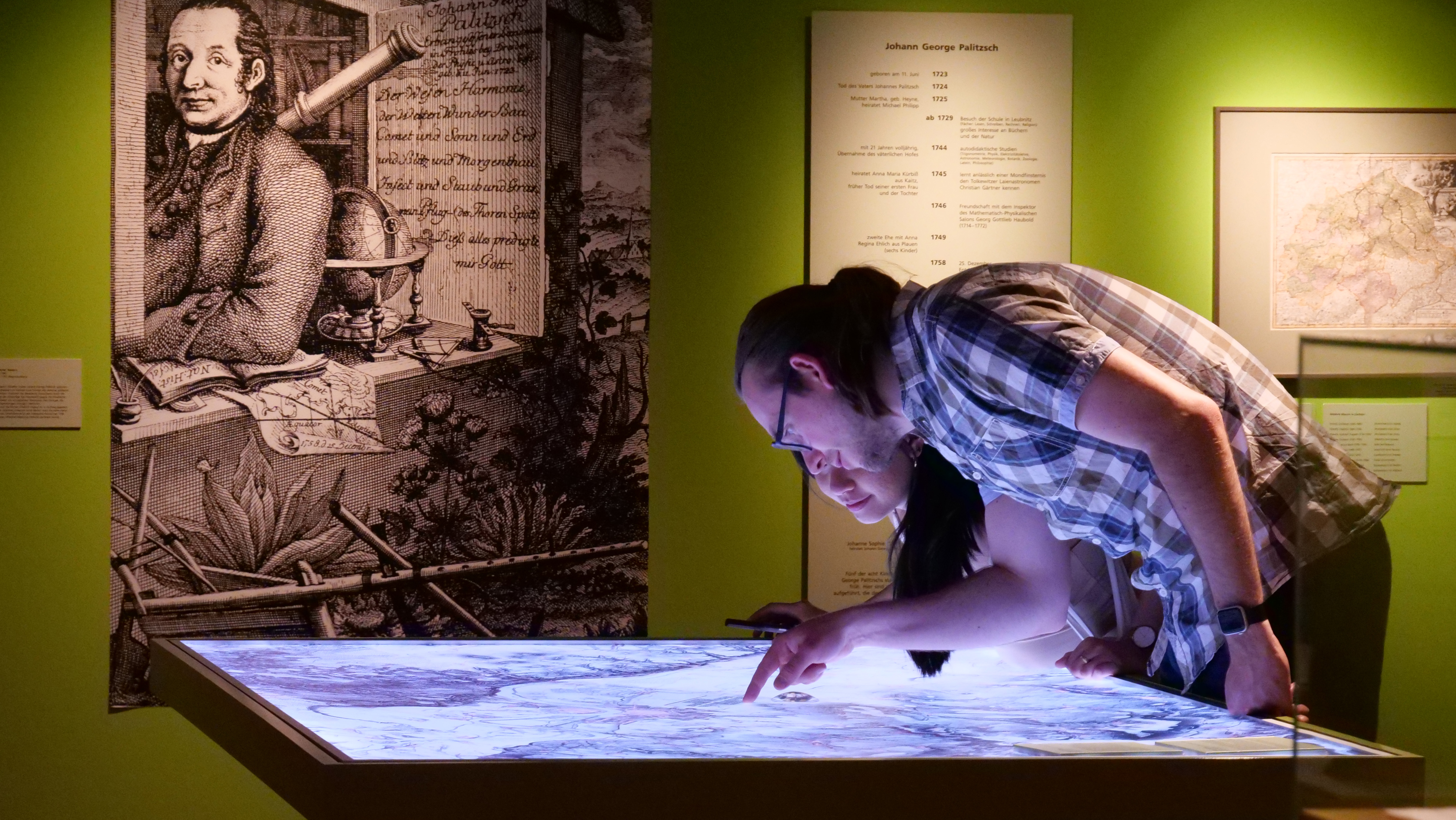 KI generiert: Zwei Personen betrachten eine interaktive, beleuchtete Karte in einem Museum oder Ausstellungsraum. Im Hintergrund ist ein historisches Porträt und eine Informationstafel sichtbar.