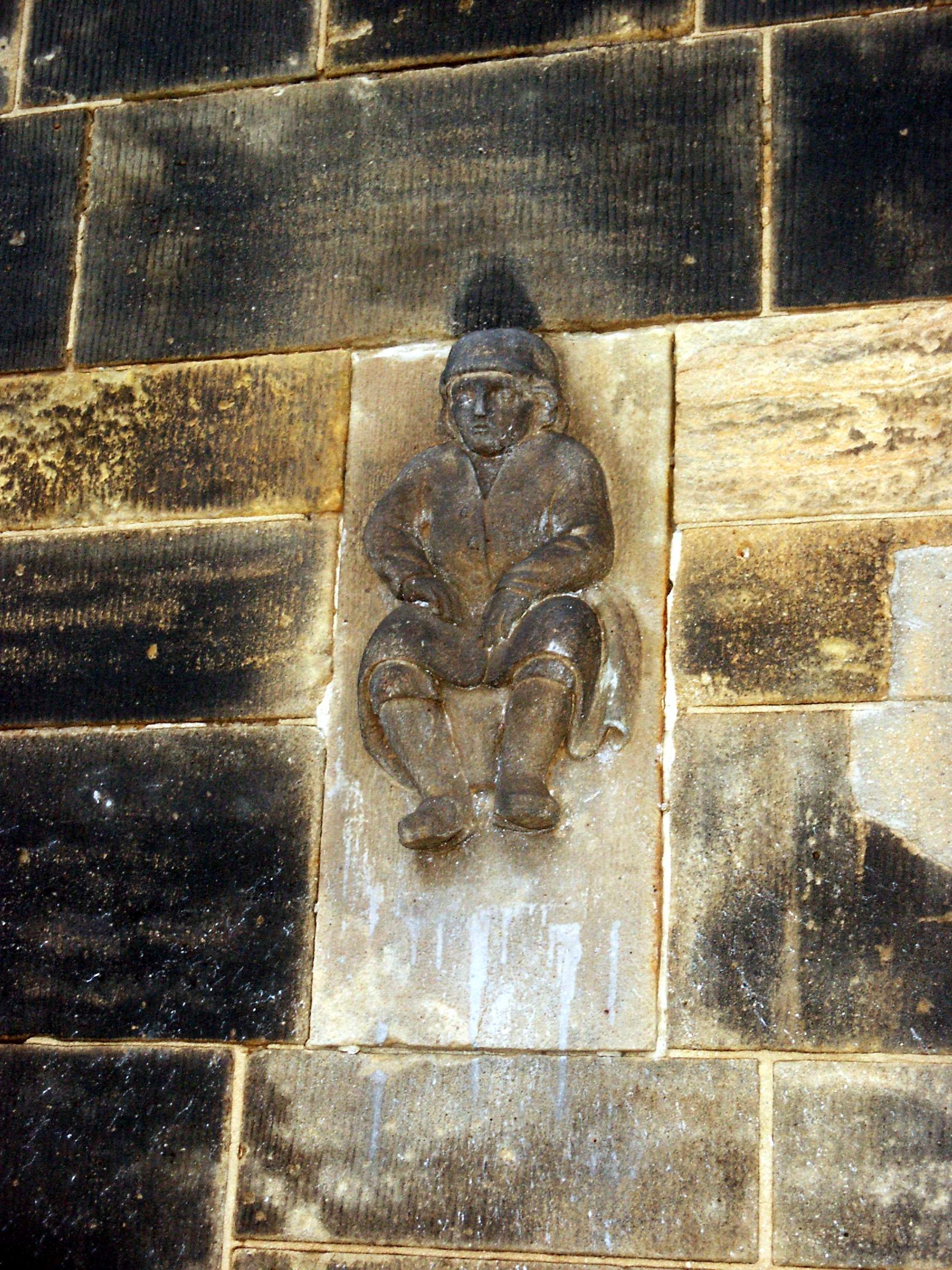 KI generiert: Das Bild zeigt eine in Stein gemeißelte Figur eines sitzenden Mannes an einer Gebäudewand. Der Hauptinhalt ist die Skulptur des Mannes, die in die Wand eingelassen ist.