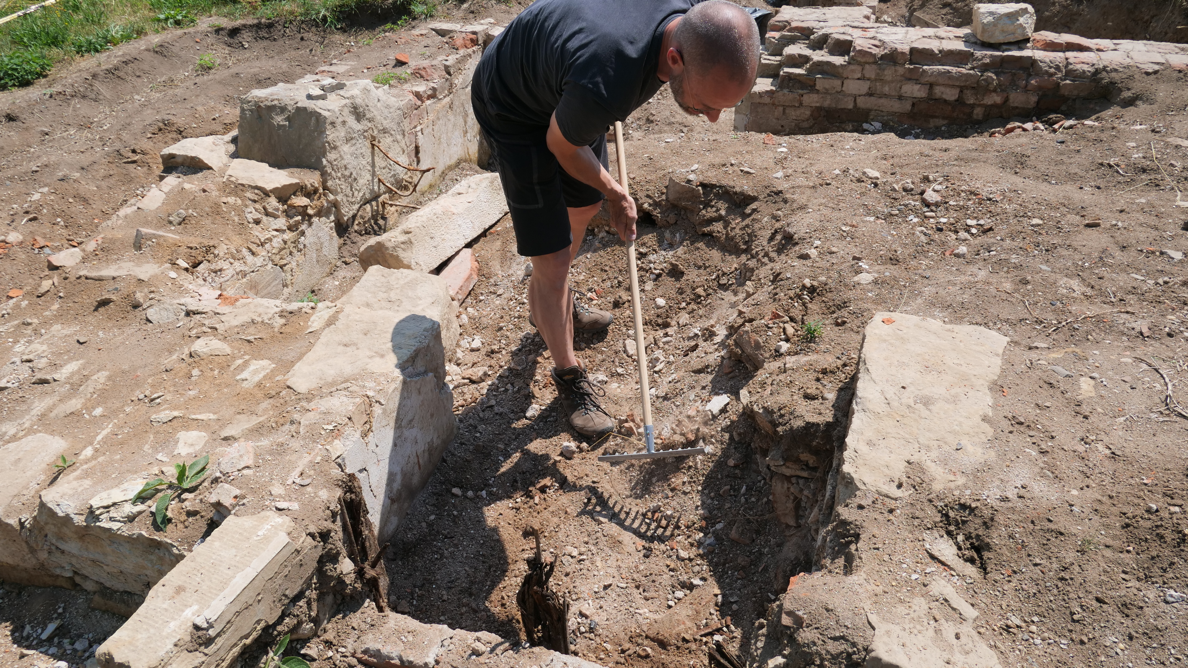 KI generiert: Das Bild zeigt eine Person, die auf einer archäologischen Ausgrabungsstelle mit einer Harke arbeitet. Der Hauptinhalt des Bildes ist die archäologische Aktivität und die freigelegten Baureste.